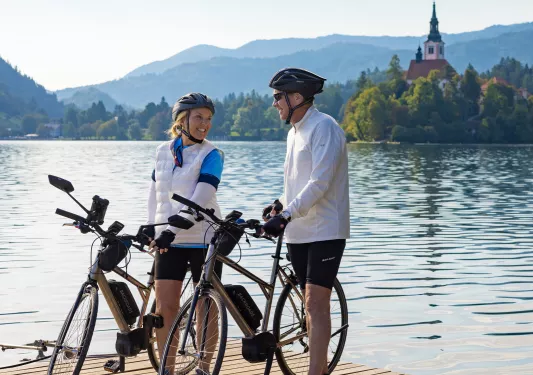 Man and woman standing next to their bikes, on a wooden bridge with a lake in the background