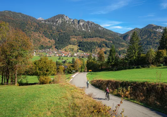 Two bikers on a road going towards a town and large mountains
