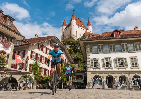 Two people riding bikes in a town square, with large buildings and stores in the background