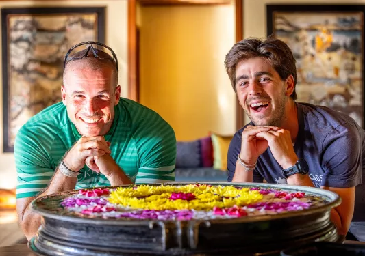 Two men smiling with standing in front of a small fountain full of flowers