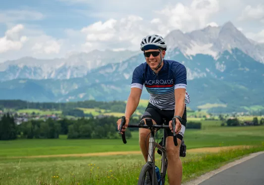 Man riding a bike on an empty road, with a large field and tall mountains in the background