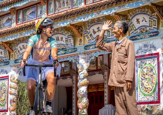 Woman riding a bike in front of a large temple, with a man smiling and waving at her