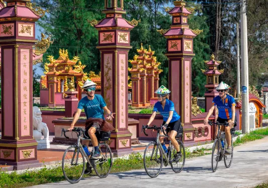 Man and two women wearing straw hats while biking next to large Vietnamese temples