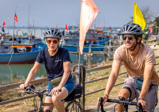 Man and woman riding bikes on a road next to a dock, with floating boats in the water