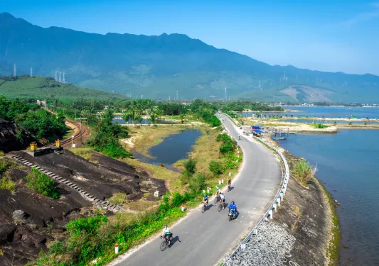 Sky view of group of people biking on an empty road, with a lake to the right and large mountain in the background