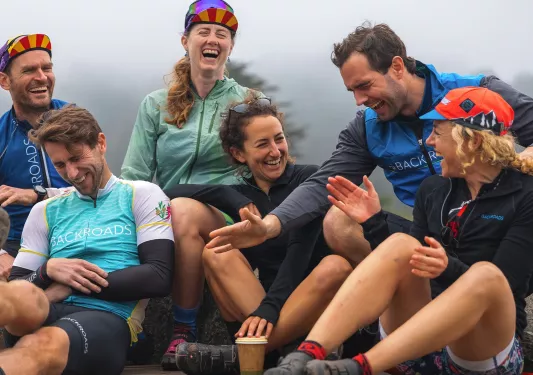 Group of people wearing biking gear, laughing while sitting on a ledge
