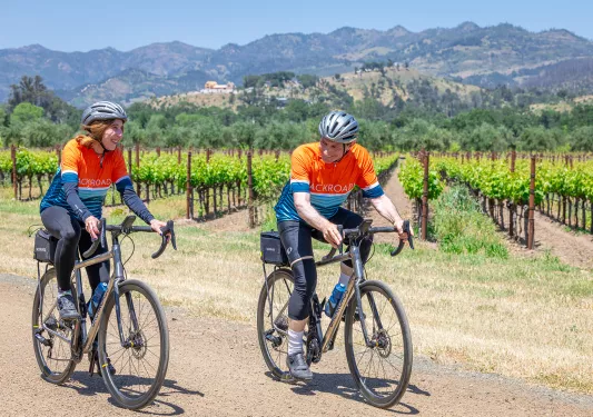Man and woman wearing orange biking gear, riding bikes in front of a field of crops