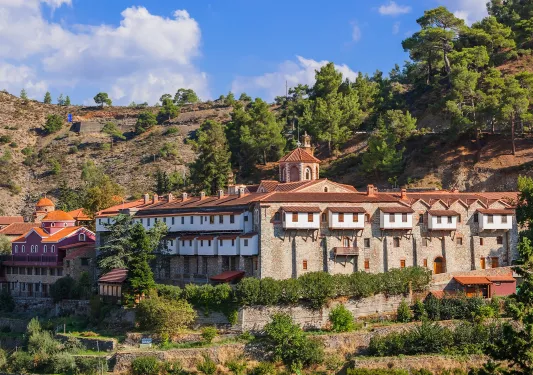 Exterior view of a stone building with red roofing, with large hills and trees behind the building