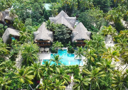 Sky view of hut-style buildings with an outdoor pool in front, surrounded by palm trees