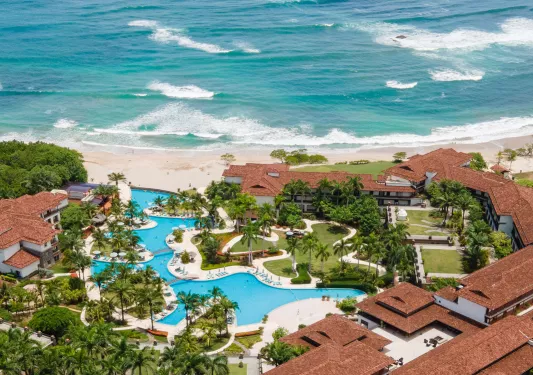 Sky view of hotel complex with an outdoor pool, with the beach in the distance