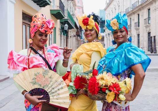 Three women in traditional dresses, holding flowers and cigars