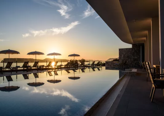 Outdoor pool, surrounded by reclining chairs and umbrellas, with the sunset in the distance