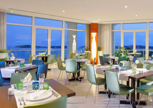 Dining hall with green chairs and wooden tables, with large windows along the walls