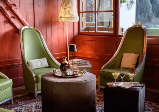 Room with red, wooden walls with green cushioned chairs and an ottoman with a bottle of liquor on top