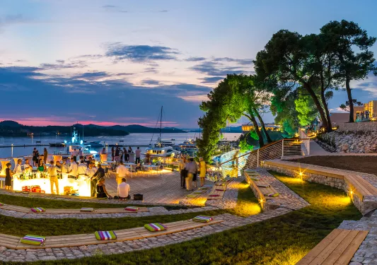 Outdoor patio and seating area with groups of people around a bar, with boats on the lake