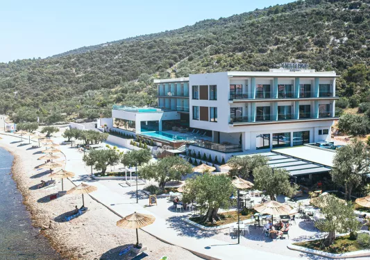 Exterior view of large, white hotel building with outdoor pools and a large forest in the back