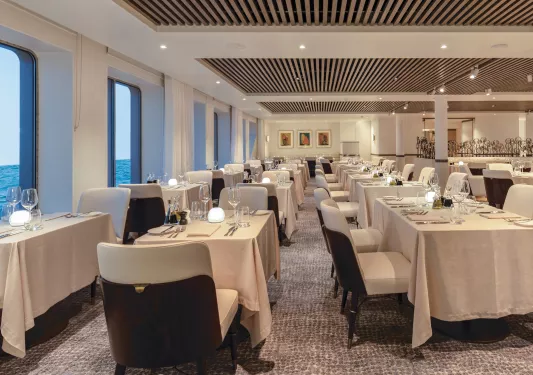 Dining hall with linen over tables and white chairs, with windows to the left looking out towards the ocean