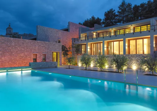 Night time view of an outdoor pool, with an illuminated building with large, glass walls and mini balconies