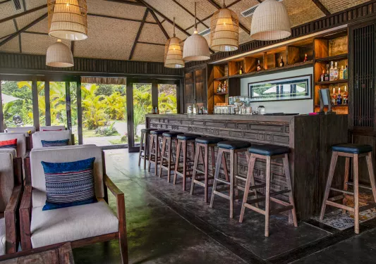 Indoor bar with a wooden counter and stools, with cushioned chairs in the background