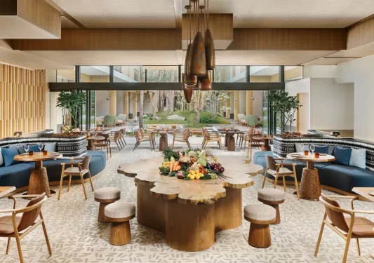 Dining hall with large wooden tables and cushioned chairs, with an outdoor garden in the background