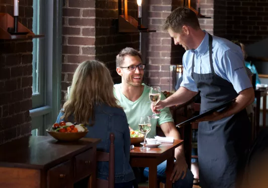 Man and woman sitting at a dining table, with a waiter handing them glasses of wine