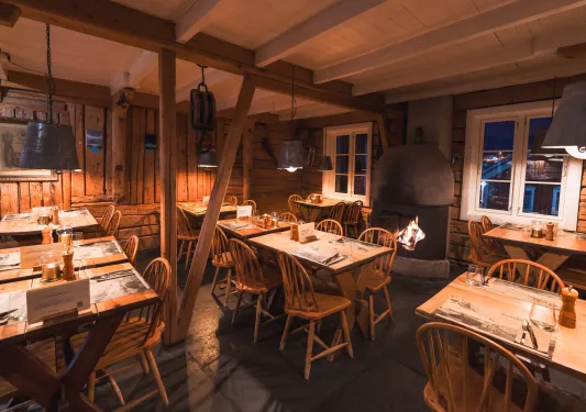 Dimly lit cabin restaurant with dining tables and wooden chairs, with a fireplace in the back