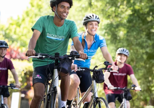 Group of men and women smiling while riding bikes on a road surrounded by tall trees
