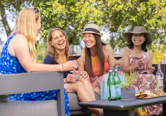 Four women outdoors, smiling and holding glasses of wine, with trees in the background
