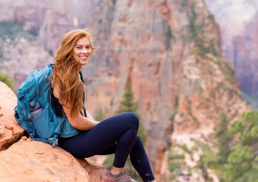 Woman wearing a blue backpack, sitting on an orange boulder with large mountains in the background