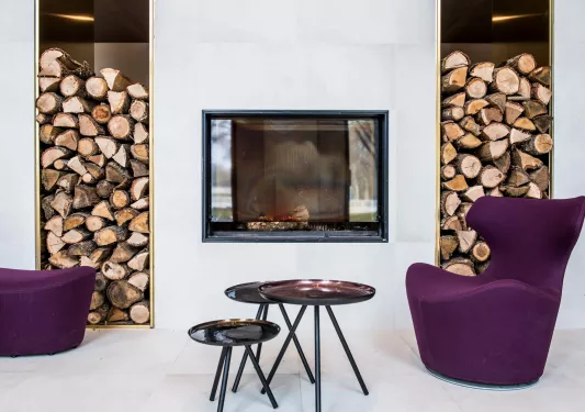 Seating area with purple, cushioned chairs, black circular tables and a fireplace in the background