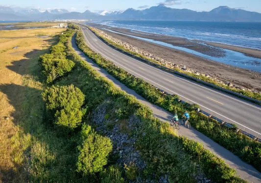 Two people riding bikes on a long trail surrounded by bushes, with a road and ocean to the right