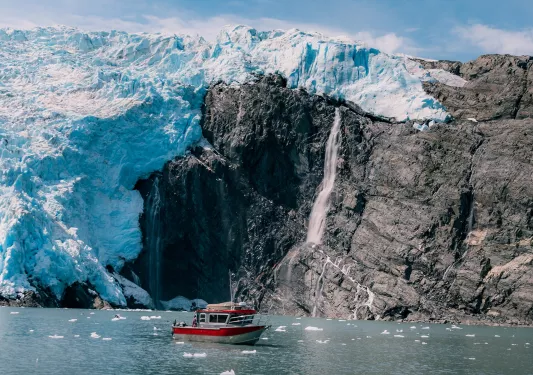 Red and gray boat floating next to large glaciers and cliffs