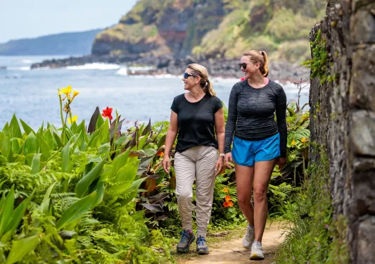 Two women smiling while walking on a dirt trail surrounded by large plants
