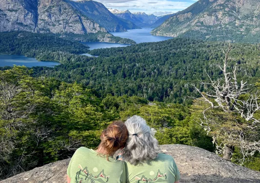 Two women wearing green shirts, sitting on a boulder looking out to a large valley of trees