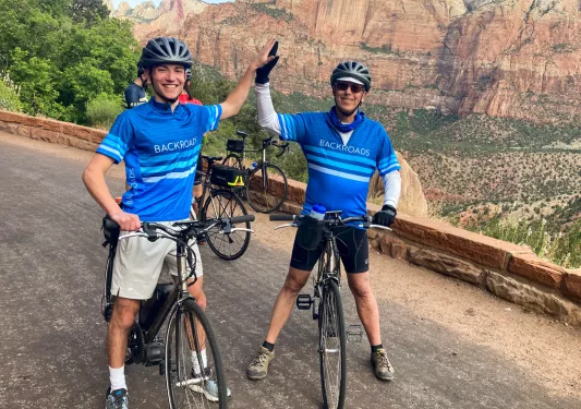 Two men wearing Backroads jerseys, high fiving each other and standing on their bikes