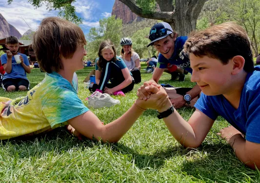 Two kids laying down on the grass, arm-wrestling