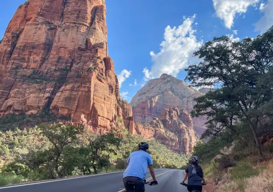 Two people biking on an empty road, surrounded by trees and tall canyons