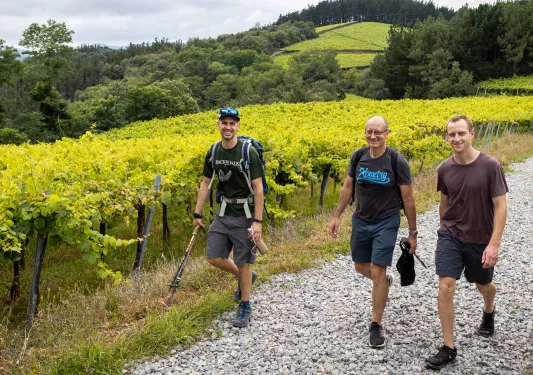 Three men smiling while hiking on a gravel trail, with crop fields in the background