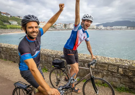 Two men biking on a path with their arms in the air, with a lake in the background