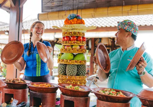 Woman and man smiling while holding lids to pots, with a tower of fruit between them
