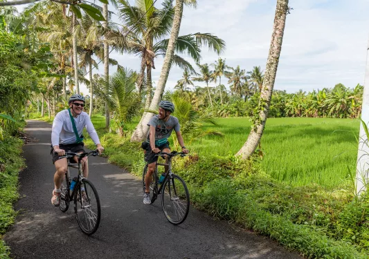 Two men riding bikes on an empty road with a large grass field and palm trees