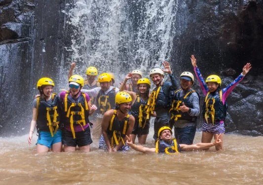 Group of people smiling while wearing helmets and life vests under a waterfall