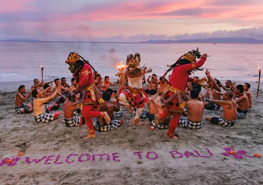 Traditional dancers on the beach, with a sign made of rose petals saying "Welcome to Bali"