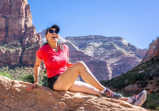 Woman wearing a pink Backroads shirt, sitting on a large boulder with mountains in the background