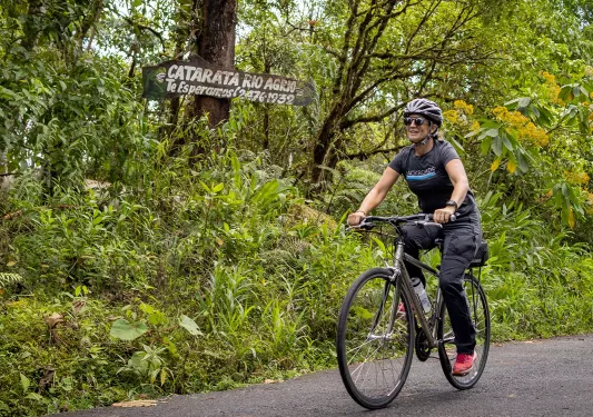 Woman biking on a road, surrounded by tall plants and trees