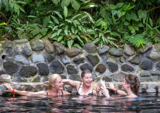 Three women holding drinks and swimming in a pool, surrounded by stone walls