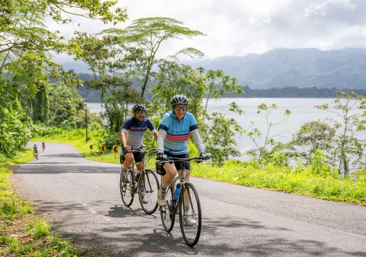 Two people riding bikes on a road, with a lake to the right
