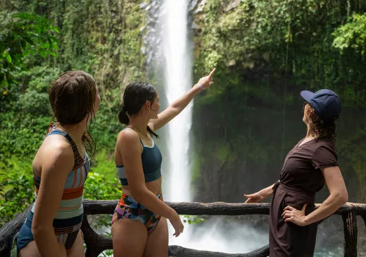 Three women standing in a forest, pointing and looking at a waterfall