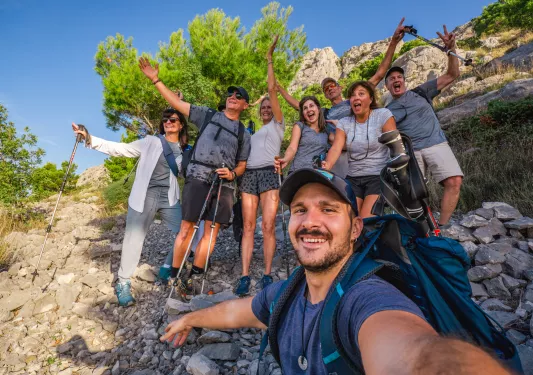 Group of men and women, holding hiking poles and smiling with their arms open on a rocky trail