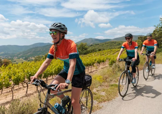 Two women and one man riding bikes on a road, with crop fields in the background
